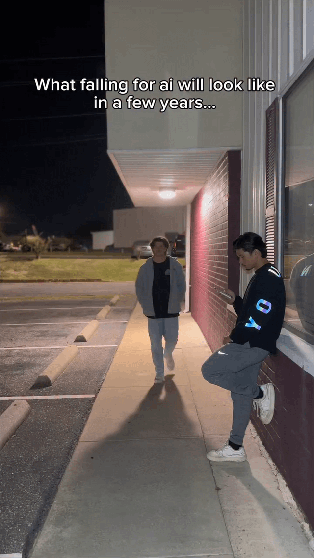 The image depicts a nighttime outdoor scene on a sidewalk adjacent to a building with a red brick wall and a lit awning. On the right side, a person is leaning against the wall, wearing a dark jacket with 'JOY' in blue letters on the sleeve, gray pants, and white sneakers, while intently looking at a mobile phone. On the left, another person, dressed in a light gray jacket, dark shirt, and light gray pants, is walking towards the camera from under the awning, which is illuminated by a ceiling light. The background shows a parking lot with a few cars and a grassy area under a dark sky. The top of the image contains the text: 'What falling for ai will look like in a few years...', suggesting a satirical commentary on potential future societal behavior where individuals may be overly engrossed in AI interactions via technology, even in social settings.