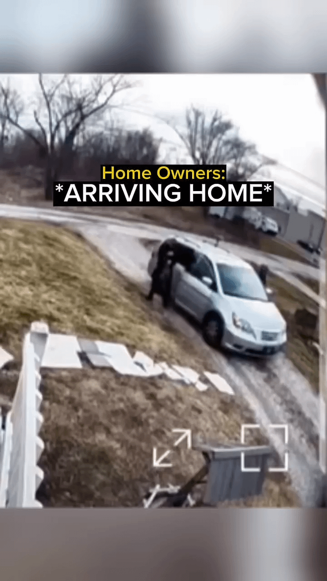 A photograph capturing a scene of home owners arriving home. A white sedan is parked on a sloped, partially snow-covered driveway with two individuals exiting the vehicle—one from the front passenger side. The background features leafless trees, a grassy embankment, and distant buildings. Overlaid text in yellow and white reads: 'Home Owners: *ARRIVING HOME*', framing the context of the moment. The image has a security camera-like quality with visible on-screen icons (arrows and a square) at the bottom, suggesting it might be a screenshot from a security feed or similar recording.