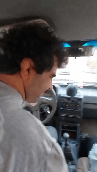 This is a low-resolution candid photograph taken from the backseat of a vehicle, depicting a man with dark curly hair wearing a light gray shirt, seated in the driver's seat facing forward. A forced perspective optical illusion is created by the camera angle: the upper curve of the steering wheel aligns perfectly with the man's facial profile, making it appear as if the steering wheel is an oversized, oddly shaped nose or mustache attached to his face. The car's interior, including the dashboard, manual gear shift, rearview mirror, and the road visible through the front windshield appear in the background. The original title "Wait for it..." references the delayed moment when viewers notice the humorous visual illusion.
