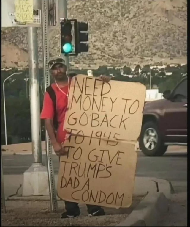 A man stands on a street sidewalk next to an active green traffic light, holding a large handwritten cardboard sign. The sign contains a dark satirical joke targeting former US president Donald Trump: it states the man needs money to time travel to 1945 to give Trump's father a condom, implying that Trump should never have been born. The background features a rocky hilly landscape, a public road with a maroon SUV driving past, and standard street infrastructure. The joke relies on crude, anti-Trump political commentary for humor.