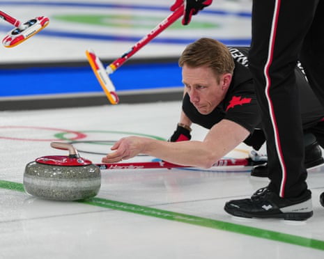 This image captures a male professional curling athlete, likely representing Canada (evidenced by the red maple leaf on his black uniform), in the middle of a stone delivery on a competitive curling rink. The athlete is crouched low to the ice, one hand guiding the grey curling stone (branded with 'Hardline') forward, with an intense, focused expression, showing full concentration on his task. In the background, curling brooms held by teammates are visible, positioned to sweep the ice once the stone is released. The rink surface features the marked target circles (the 'house') and boundary lines, confirming a formal competitive setting, such as an Olympic or international curling championship.