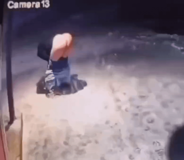 A grainy security camera still showing a person bending over in an outdoor paved area. The person appears to be wearing a light-colored top and dark pants. The environment is dimly lit with shadows visible on the ground. The text 'Camera13' is displayed in the top-left corner, indicating this is from the 13th security camera in a system.