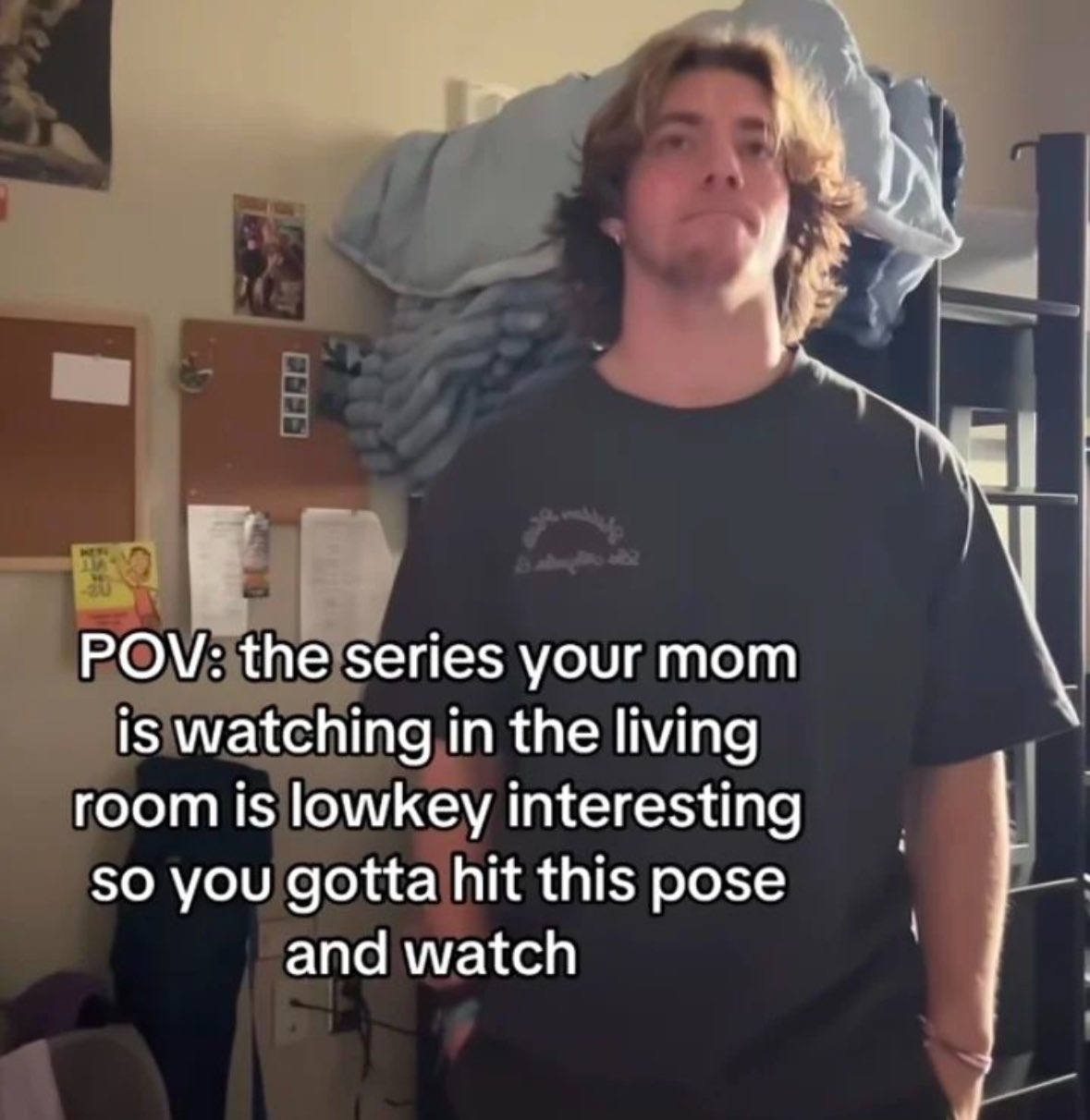 The image is a single photograph of a young Caucasian man with wavy, shoulder-length blondish-brown hair, wearing a dark t-shirt, standing in a casual indoor space that appears to be a dorm room or bedroom, with bunk beds, cork boards, and wall decorations visible in the background. He is holding a deliberately nonchalant, uninterested expression, with his hands in his pockets, looking slightly upward and off-camera. Overlaid white text on the left side of the image reads 'POV: the series your mom is watching in the living room is lowkey interesting so you gotta hit this pose and watch'. The meme jokes about the common relatable experience of being unexpectedly drawn into a television series that a parent is watching, but choosing to stand nearby feigning disinterest rather than admitting you want to watch the show.