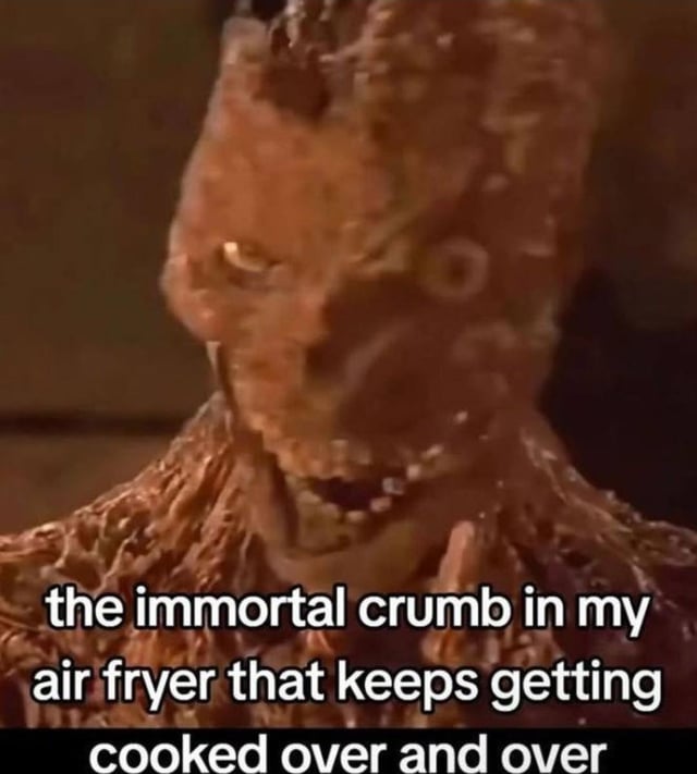A meme featuring a close-up of a grotesque, menacing monster with rough, textured skin, glowing yellow eyes, and a snarling mouth. Overlaid text at the bottom reads: 'the immortal crumb in my air fryer that keeps getting cooked over and over'. The monster's intimidating appearance humorously contrasts with the mundane scenario of a persistent, hard-to-remove crumb in an air fryer, creating a relatable joke about everyday household frustrations with kitchen appliances.