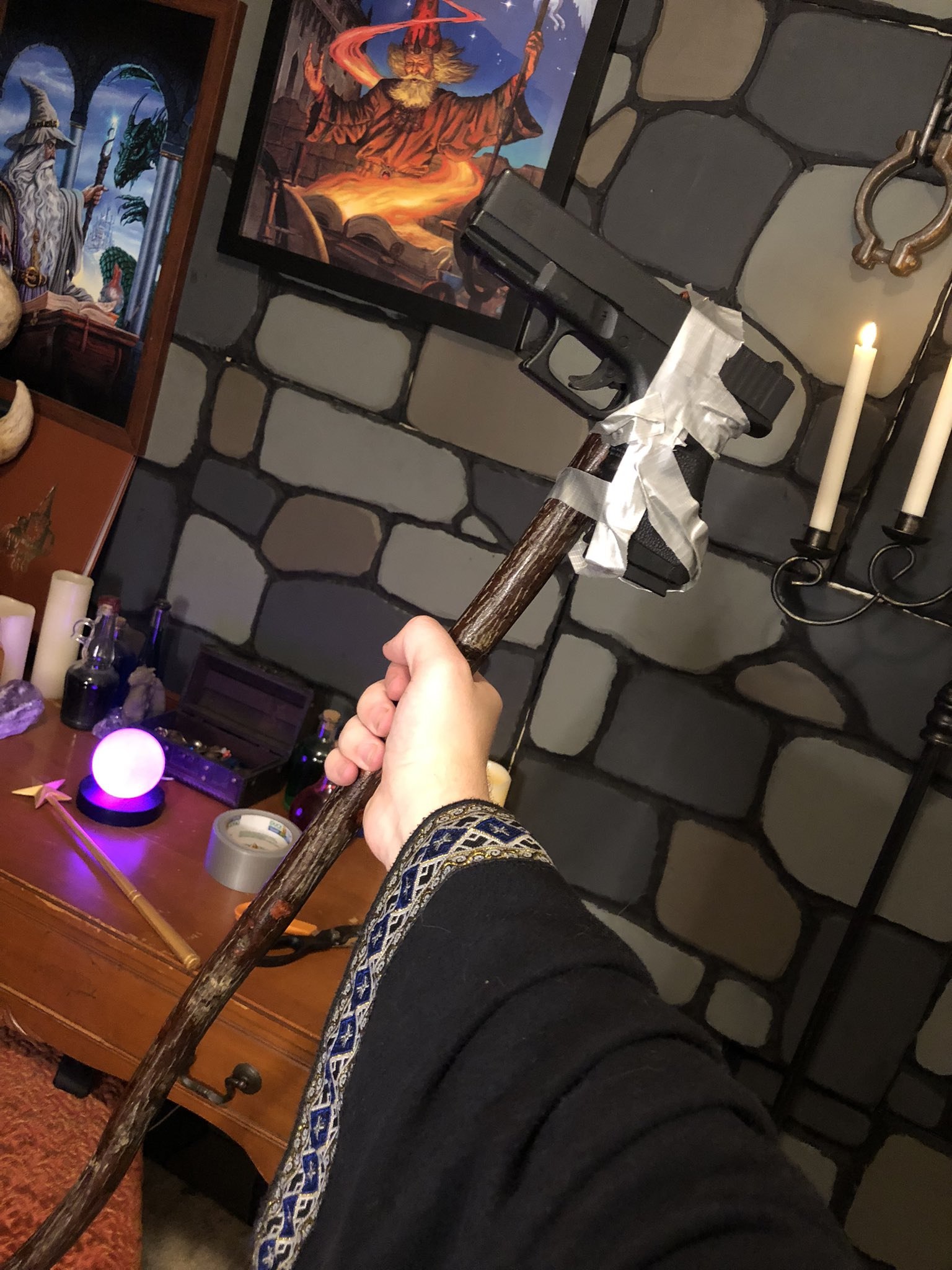 A first-person perspective photograph showing a hand holding a rustic wooden staff with a black handgun crudely taped to its top. The background features a stone-textured wall adorned with framed fantasy artwork depicting wizards and magical scenes. A wooden desk in the foreground holds various magical-themed items: a glowing purple crystal ball, a small treasure chest, glass bottles, a lit candle, and a roll of silver tape (likely used to attach the gun to the staff). The person’s sleeve is black with intricate blue and gold embroidery, enhancing the fantasy aesthetic. The image humorously blends medieval magical elements with modern weaponry, creating a whimsical DIY creation.