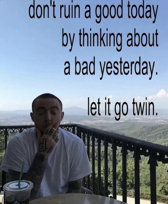 A photograph of a real person sitting at an outdoor table with a scenic backdrop of mountains, trees, and a distant town. The individual is wearing a white t-shirt, has visible tattoos on their forearm, and is positioned with one hand near their mouth, suggesting a relaxed or contemplative pose. A clear plastic cup with a green straw (resembling a Starbucks cup) sits on the table in front of them. Overlaid text in black font reads: 'don't ruin a good today by thinking about a bad yesterday. let it go twin.' The image combines a casual, relatable setting with motivational messaging to encourage releasing past negativity.