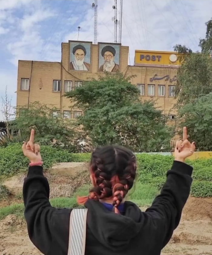A photograph capturing the rear view of a person with braided hair, wearing a black hoodie, standing in front of a multi-story brick building. The individual raises both hands with middle fingers extended upwards, directed towards the building. The building features two large portraits of bearded men on its upper facade, a yellow sign with the word 'POST' in uppercase letters, and non-English text (likely Persian) on the right side. The scene includes greenery, bushes, and trees in the foreground, with the person positioned on a grassy and dirt area.