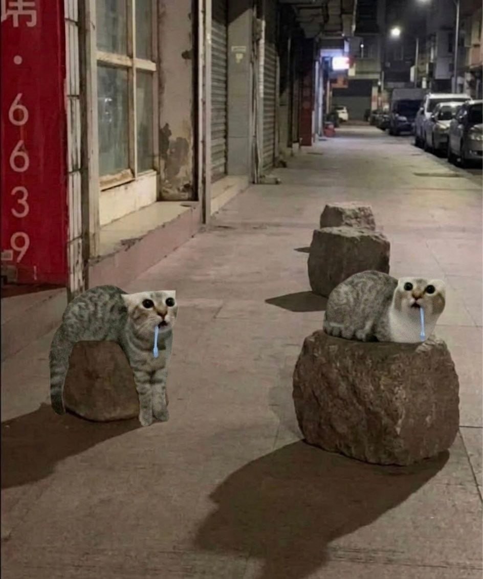 The image captures a dimly lit urban sidewalk at night, with closed shuttered storefronts on the left and parked cars lining the right side of the street. Four rough, unrefined stone bollards are spaced out along the sidewalk. The two front bollards have been digitally edited to have the upper half of the popular wide-eyed, drooling gray tabby cat meme emerging from the top of each stone, making the cats appear as if they are partially made of stone, or sitting perched on the bollards with identical vacant, dribbling expressions. The two back bollards remain unedited, creating a humorous progression effect.