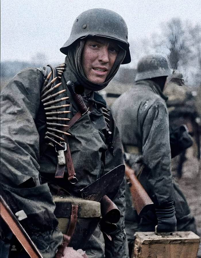 This is a colorized historical photograph capturing a German infantry soldier from World War II in a muddy, cold frontline environment. The foreground soldier wears a weathered M1935 Stahlhelm (steel helmet) with a cloth liner, a tattered field gray uniform, and a bandolier of rifle ammunition draped across his chest. He has a weary, strained but alert expression, looking directly at the camera. His combat gear includes a field canteen, a sheathed combat knife, and part of his rifle is visible at his side. In the background, another soldier with his back turned stands near a smoking area, suggesting a temporary field rest stop, with bare trees and an overcast sky emphasizing the harsh, cold conditions of the combat theater. The image vividly conveys the gritty, exhausting reality of frontline military service during the war.
