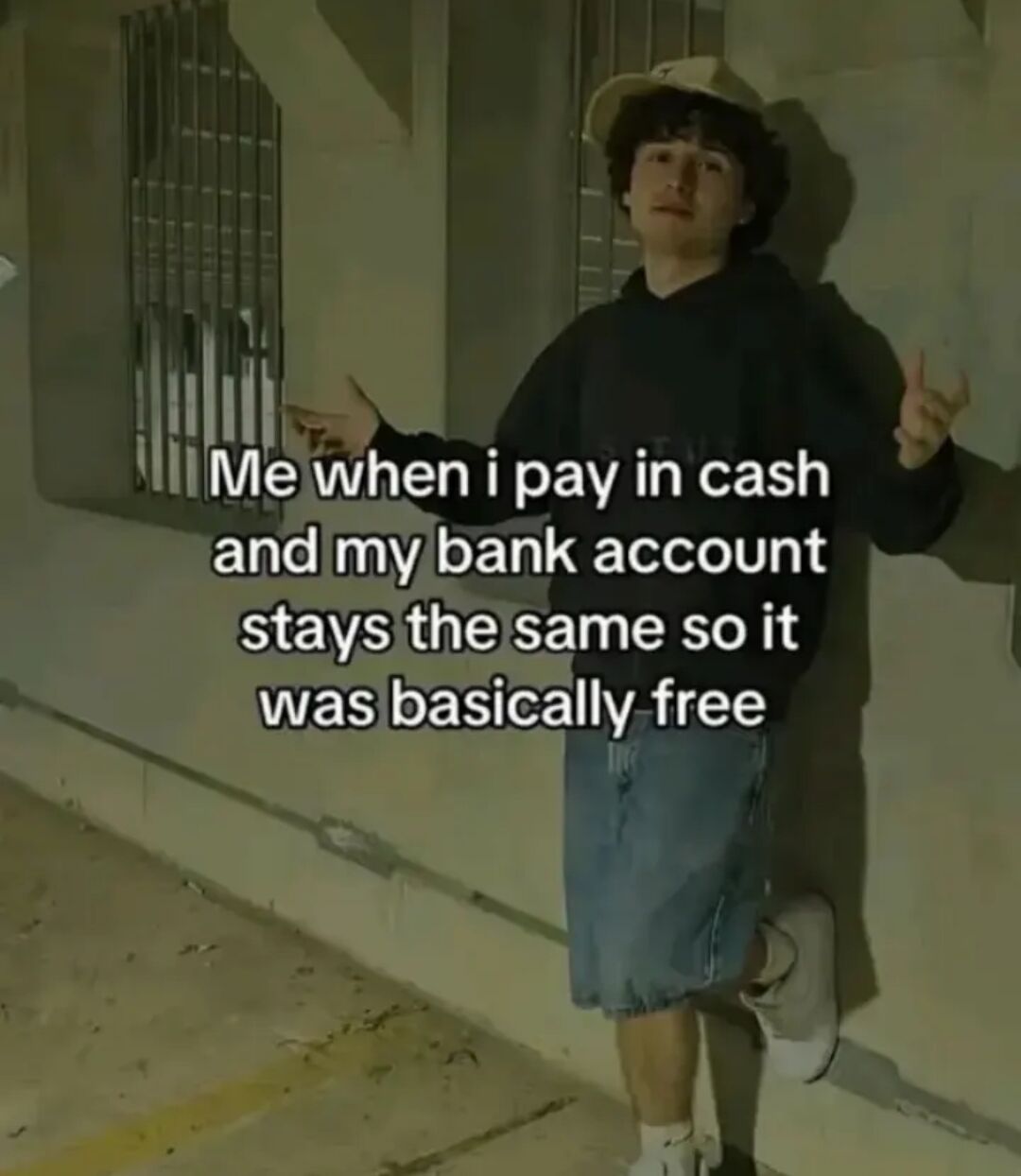 This is a relatable humor meme featuring a real young man leaning against a concrete industrial-style building wall in an outdoor alley setting, dressed casually in a beige bucket hat, black hoodie, baggy knee-length denim shorts, and white sneakers. He is posed with both hands held out in a casual, unbothered, justifying gesture, one foot propped against the wall. Overlaid white text on the image describes a common silly flawed logic: 'Me when i pay in cash and my bank account stays the same so it was basically free', poking fun at how people often mentally separate physical cash spending from their bank account balance, leading them to pretend cash purchases cost them nothing.