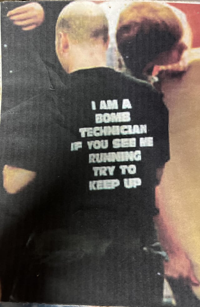 A photograph showing the back of a person with a bald head wearing a black shirt. The shirt features bold white text that reads: 'I AM A BOMB TECHNICIAN IF YOU SEE ME RUNNING TRY TO KEEP UP'. The individual is standing in a group setting, with part of another person (brown hair, partial profile) visible to their right. The image has a slightly grainy, low-contrast quality, suggesting it may be a scanned printed photo or a low-resolution digital capture.