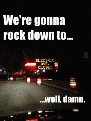 A nighttime photograph depicting a road closure scene with a neon sign reading 'ELECTRIC AVE CLOSED' in the background, flanked by traffic cones and vehicle headlights. Overlaid text at the top reads 'We're gonna rock down to...' and concludes at the bottom with '...well, damn.' The meme humorously subverts the expectation of completing the lyric from the song 'Electric Avenue' by Eddy Grant, replacing the anticipated location with a road closure, resulting in a tone of comedic disappointment.