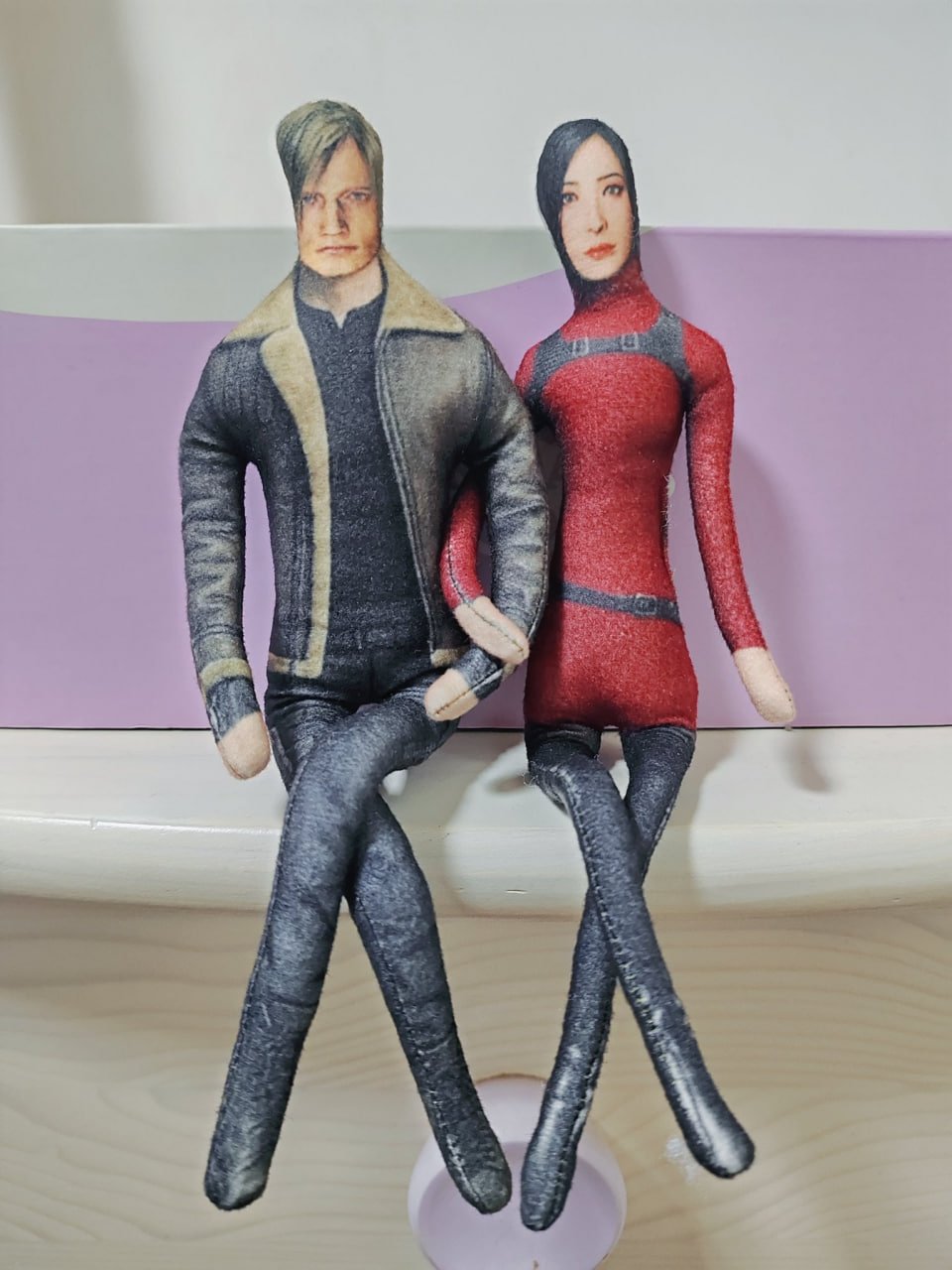 This image displays two handmade custom plush dolls of Leon S. Kennedy and Ada Wong, iconic characters from the *Resident Evil* video game franchise. Leon is outfitted in his signature brown shearling-lined jacket and dark pants, while Ada wears her recognizable form-fitting red and black bodysuit. The dolls are posed sitting side-by-side, holding hands, with their legs dangling over a white flat surface against a soft purple backdrop. The characters' faces are printed to match their in-game likenesses, while their hands are simple, unprinted felt pieces. The piece is a wholesome, fan-created take on the beloved gaming duo.