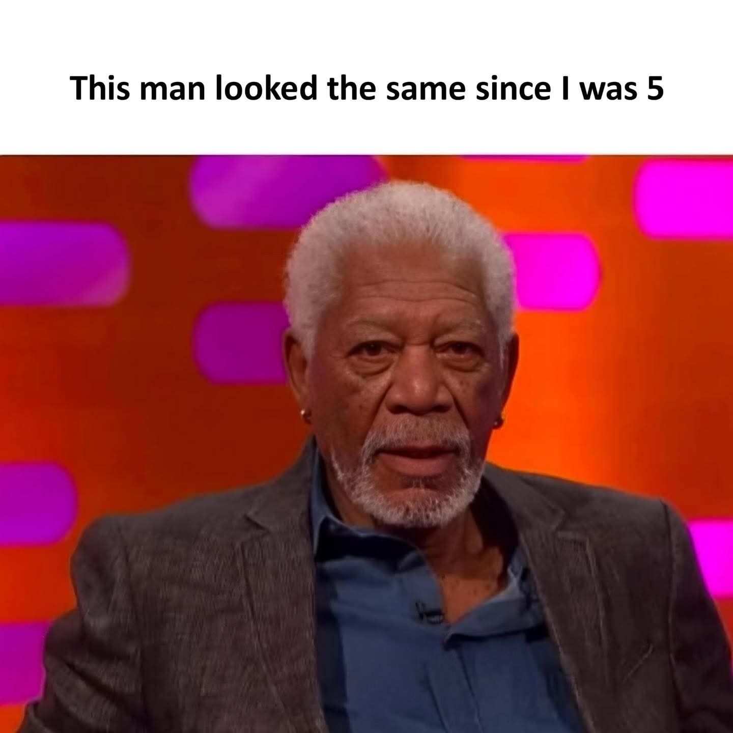This is a relatable wholesome meme featuring a headshot of well-known actor Morgan Freeman sitting on a talk show set with a vibrant orange and purple glowing background. Above the photograph, black text reads "This man looked the same since I was 5", referencing the widely shared popular observation that Morgan Freeman has appeared to age extremely slowly for many decades, remaining visually consistent across multiple generations of viewers.