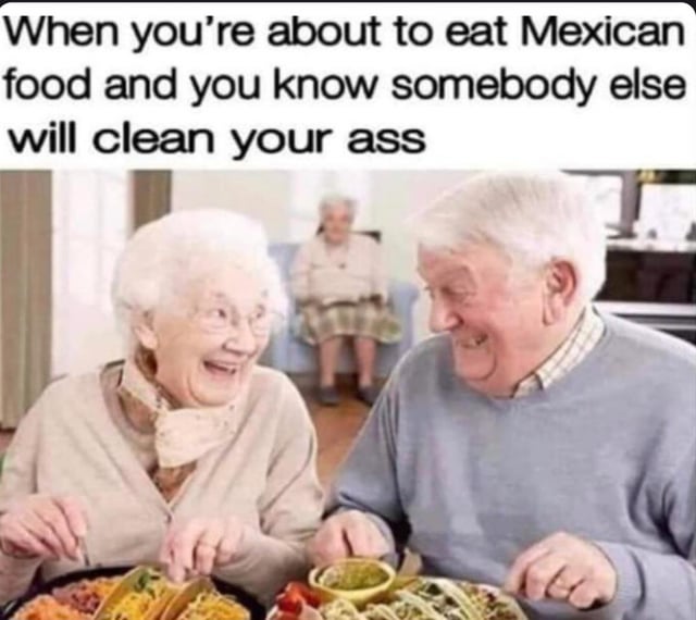 The meme features a photograph of two elderly individuals (a man and a woman) sitting at a table with plates of Mexican food, including tacos and a bowl of salsa. Both are laughing heartily, with the woman on the left wearing a light-colored sweater and scarf, and the man on the right in a gray sweater. In the background, another elderly person is visible sitting on a couch. The text overlay at the top reads: 'When you're about to eat Mexican food and you know somebody else will clean your ass.' The humor stems from the double entendre of 'clean your ass'—referring to both cleaning up the mess from eating and playfully alluding to potential digestive effects of Mexican food, though the primary joke is the relief of not having to clean up oneself.