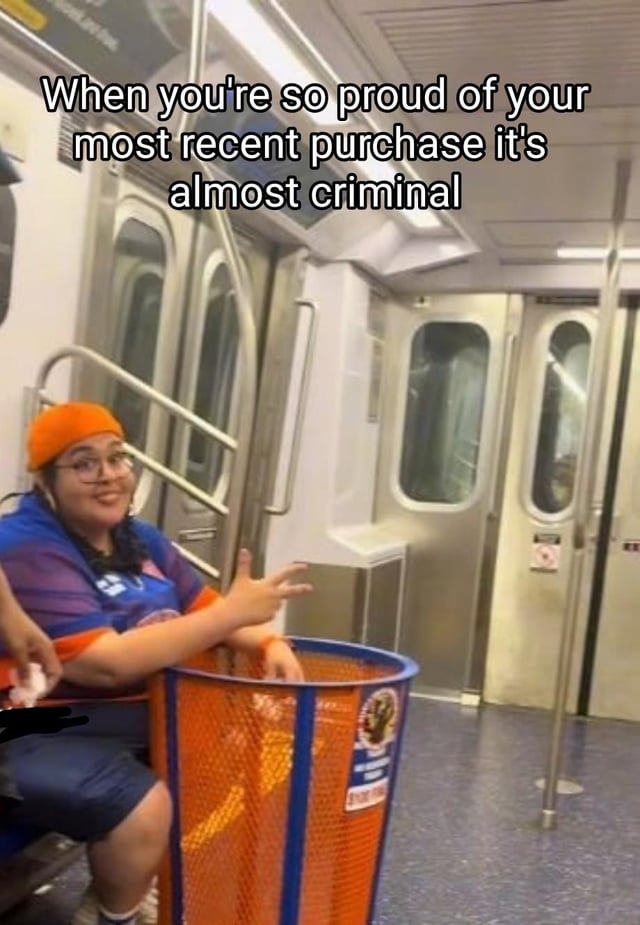 The image is taken inside a subway car. A woman wearing an orange beanie, glasses, and a blue and orange sports jersey is sitting on a subway seat, grinning proudly while gesturing at an orange and blue public trash bin placed right next to her as if it is a valuable new item she bought. Overlaid text at the top of the image reads "When you're so proud of your most recent purchase it's almost criminal". The humor comes from the absurd premise of treating a public, non-purchased trash bin like a coveted, prized new possession, leaning into relatable jokes about feeling overly excited about odd or trivial purchases.