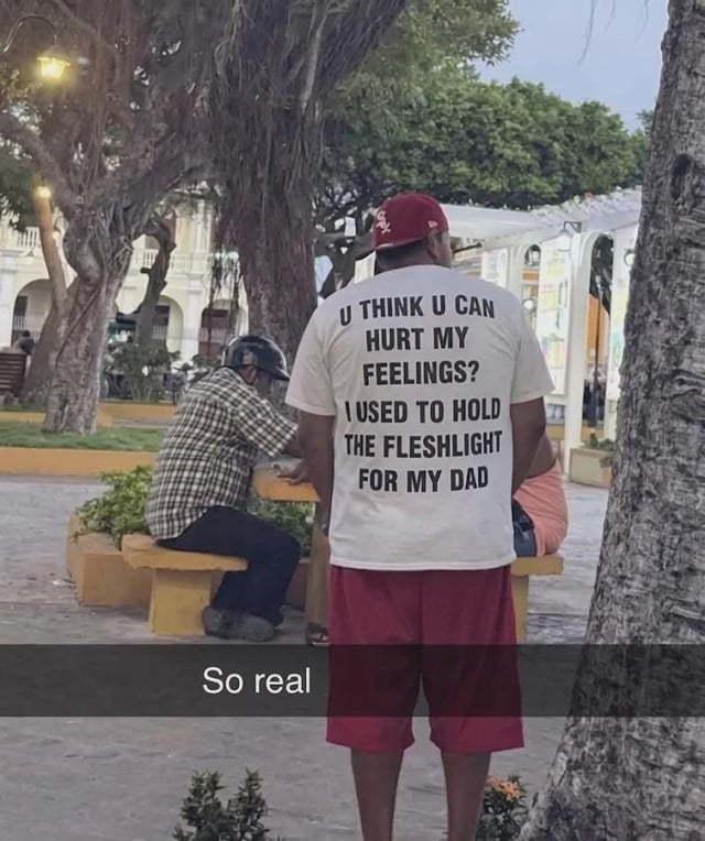 This candid meme image taken in a public outdoor park shows a man standing with his back to the camera, wearing a white custom printed t-shirt with shocking humorous text. The text on the back of the shirt reads: 'U THINK U CAN HURT MY FEELINGS? I USED TO HOLD THE FLESHLIGHT FOR MY DAD'. An overlaid caption below the image says 'So real', an internet slang reaction that emphasizes how 'relatable' the absurd text is as a joke. The humor comes from the unexpected, awkward, inappropriate implication of the statement, leaning into shock value and absurdity.