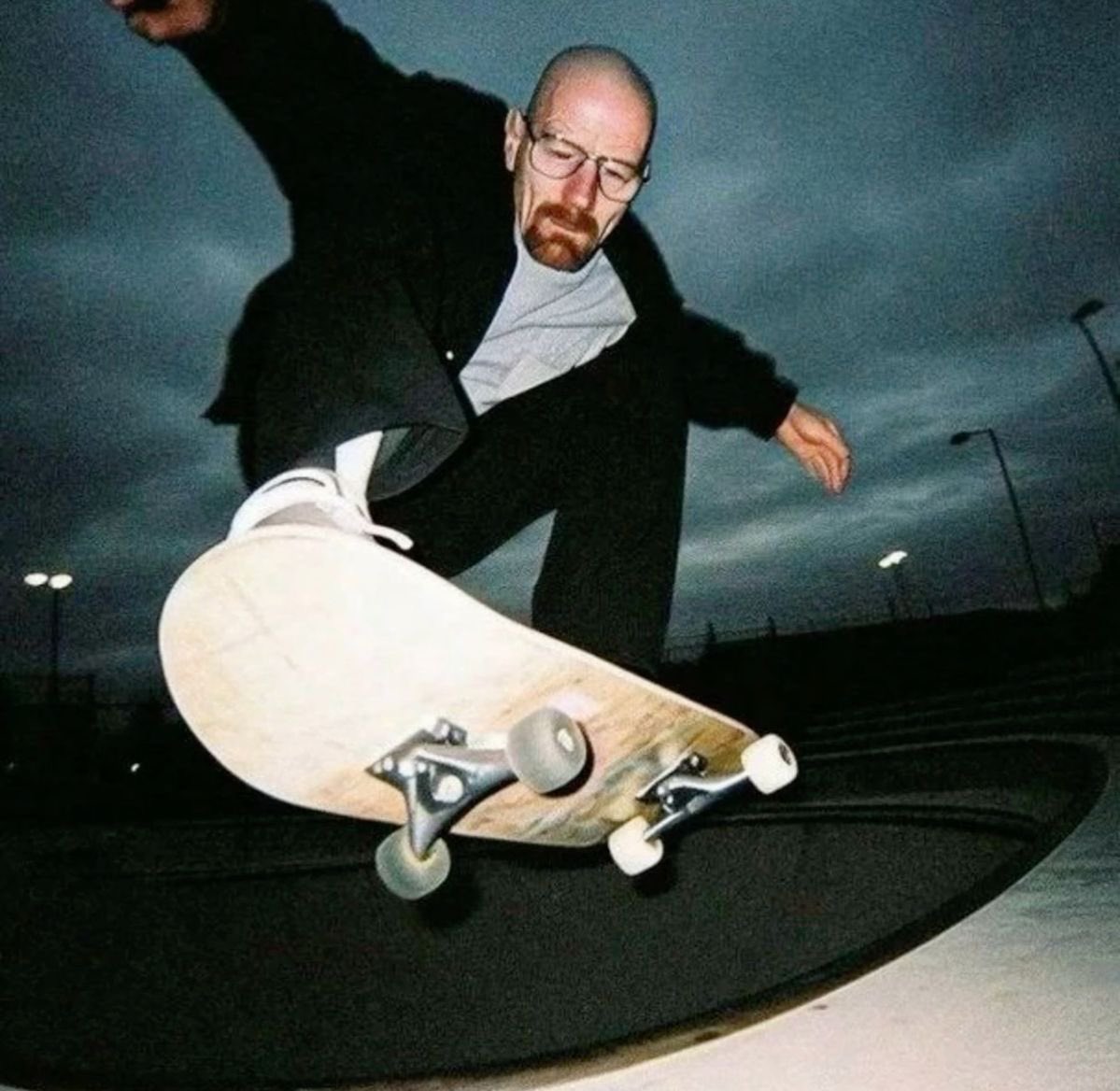 This is an edited humorous meme where the face of Walter White, the iconic protagonist of the TV series *Breaking Bad* (a bald middle-aged man with rimless glasses and a reddish goatee), is superimposed onto the body of a skateboarder performing a trick at an outdoor skatepark at dusk. The skateboarder is mid-action, leaning forward with arms out for balance, wearing a black blazer, grey undershirt, black trousers, and white sneakers, with one foot on a light-colored skateboard angled toward the camera. The background features a dark, overcast dusk sky with visible streetlights in the distance. The joke comes from the extreme incongruity of the normally serious, stoic Walter White character participating in a casual, youthful extreme sport activity that is entirely out of line with his established on-screen persona.