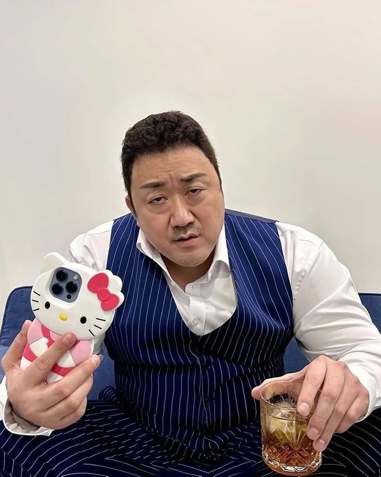 A man seated on a blue couch against a plain white background. He wears a white dress shirt under a blue vertical pinstripe vest. In his left hand, he holds a smartphone encased in a large, 3D Hello Kitty cover with a pink bow, white face, and pink body. His right hand holds a clear glass filled with a brown liquid and ice. His expression appears neutral to slightly confused, creating a humorous contrast between his formal attire and the playful phone case.