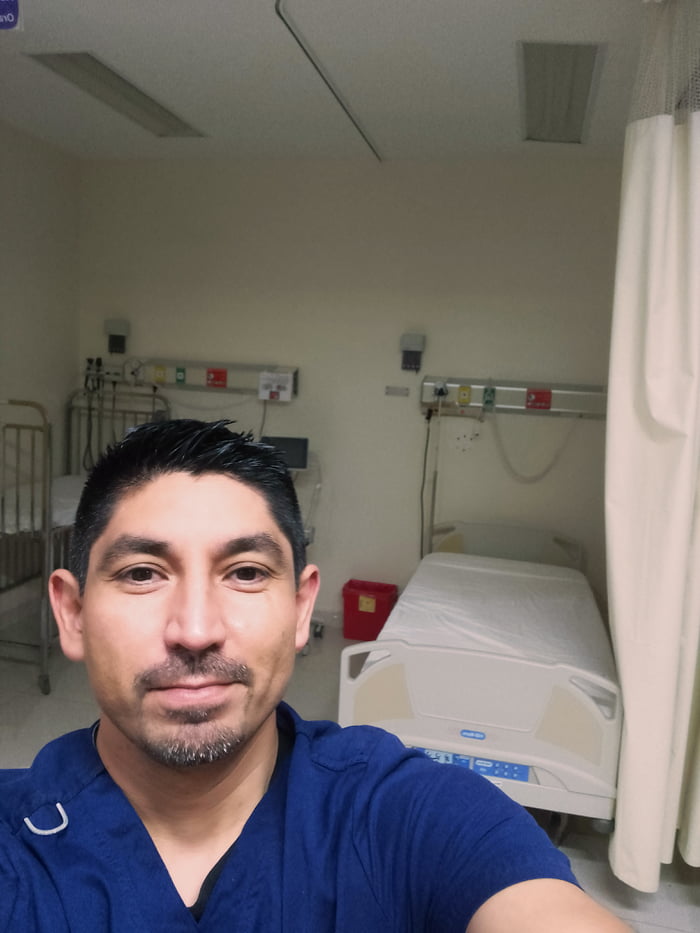 A selfie photograph of a male healthcare worker wearing blue scrubs, standing in a hospital room. The background features multiple hospital beds with white linens, medical monitoring equipment attached to the walls, a red trash bin, and a beige privacy curtain. The individual is positioned in the foreground, facing the camera with a neutral expression and a slight smile, suggesting a calm and friendly demeanor. The clinical setting is evident through the presence of IV poles, wall-mounted medical devices, and standard hospital furnishings, indicating a workplace environment during what appears to be a holiday period.