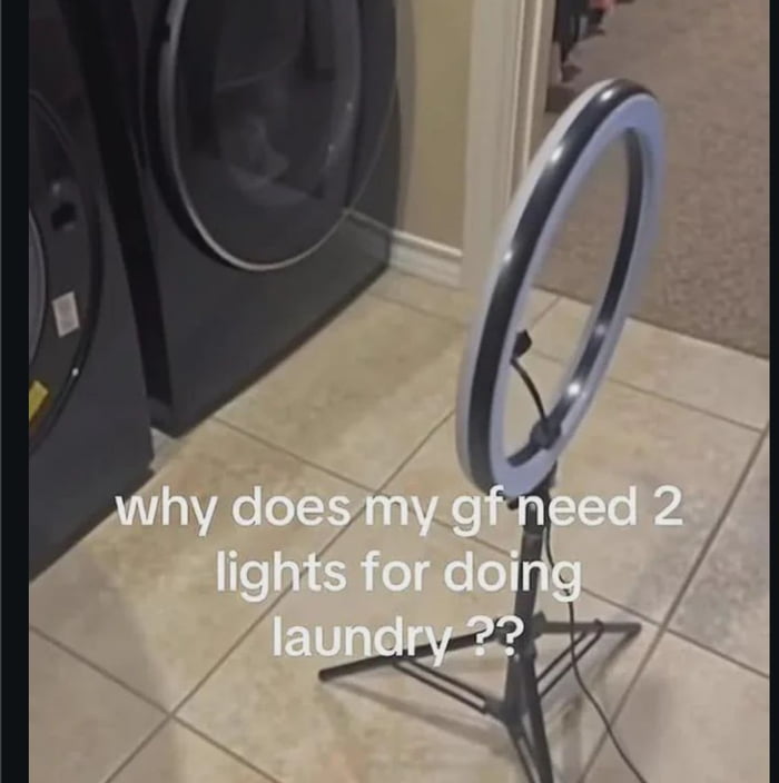 The image is shot in a home laundry area, showing a black front-loading washing machine with a round glass door next to a small standing LED ring light placed on light-colored tiled flooring. White text is overlaid on the image reading 'why does my gf need 2 lights for doing laundry??'. The joke comes from the visual similarity between the circular washing machine door and a second ring light, with the poster feigning obliviousness to the fact that his girlfriend is using the ring light to film content (such as social media videos or adult content) in the laundry room rather than just doing laundry.