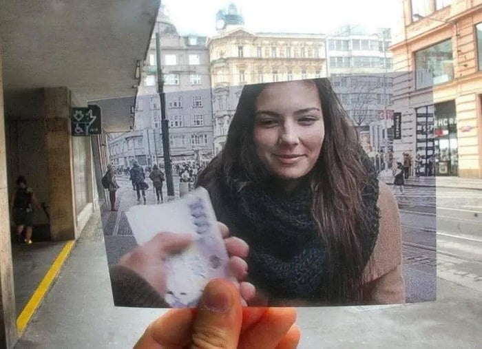 A meme utilizing a trompe-l'oeil technique where a hand holds a printed photo of a smiling woman with long dark hair and a black scarf. The photo is overlaid onto a real-world background of a busy urban street featuring classical European architecture, pedestrians, and tram tracks. The foreground shows a concrete underpass with a yellow line and a distant pedestrian, creating the illusion that the woman in the photo is physically present in the scene.