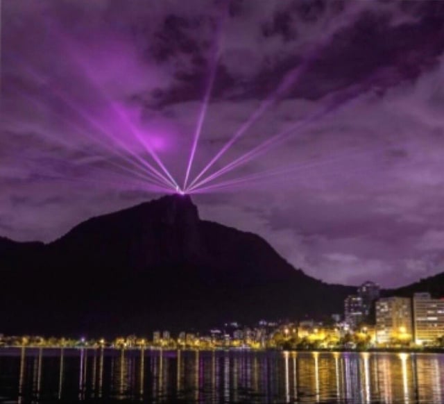 A nighttime landscape image featuring a dark, prominent mountain with bright purple laser-like beams radiating from its summit. Below the mountain, a city with illuminated buildings is visible, and the water in the foreground reflects the city lights. The purple beams resemble the visual effect that occurs when the Ender Dragon is defeated in the game Minecraft, creating a humorous connection between the real-world mountain scene and the game's iconic boss battle.