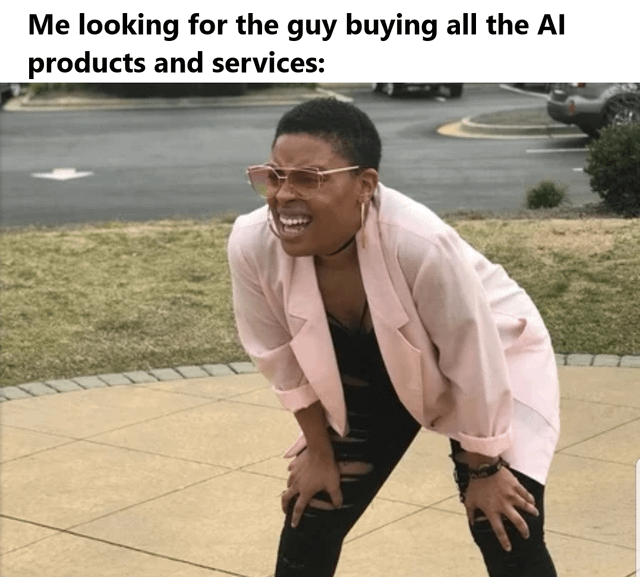A meme consisting of a top text caption and a photograph. The top text reads: 'Me looking for the guy buying all the AI products and services:'. Below the text is a photograph of a person wearing a light pink blazer, black ripped jeans, and glasses, bending forward with hands on their knees, looking around with a wide, amused smile. The background shows a paved area, grass, and a parking lot with cars, suggesting an outdoor suburban or urban setting. The image humorously portrays someone eagerly searching for an individual who is purchasing numerous AI products and services, likely commenting on the trend of AI product consumption.