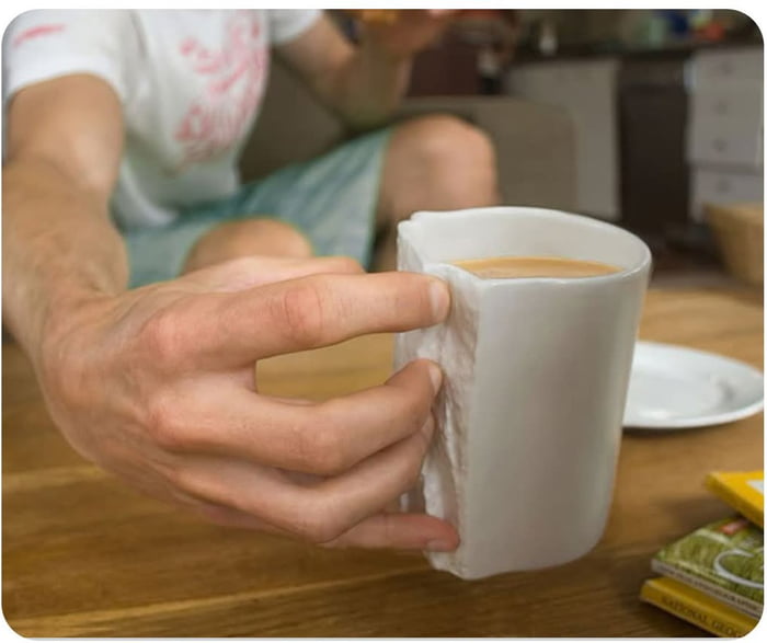 A close-up photograph of a person's hand holding a white ceramic coffee cup filled with light brown coffee. The hand is gripping the cup with a white napkin wrapped around the side, likely to insulate against heat. In the background, a wooden table is visible with a white plate and several colorful rectangular items (possibly tea or coffee packets) stacked in the lower right corner. A person is partially visible sitting in the background, wearing a white shirt with pink designs and light blue shorts, with legs bent and one hand holding an indistinct object.