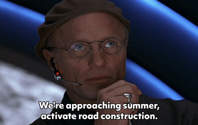 This meme uses a screenshot of Christof, the fictional showrunner from the 1998 film *The Truman Show* played by Ed Harris. He is depicted in the show's control room, wearing a communication headset, looking serious and authoritative as if issuing an official command. The overlaid text reads "We're approaching summer, activate road construction.", referencing the widely relatable, sarcastic observation that road construction projects appear en masse every summer, causing frequent, long-lasting traffic disruptions. The original post title adds the implied punchline that this construction is intentionally done as slowly as possible to extend public inconvenience.