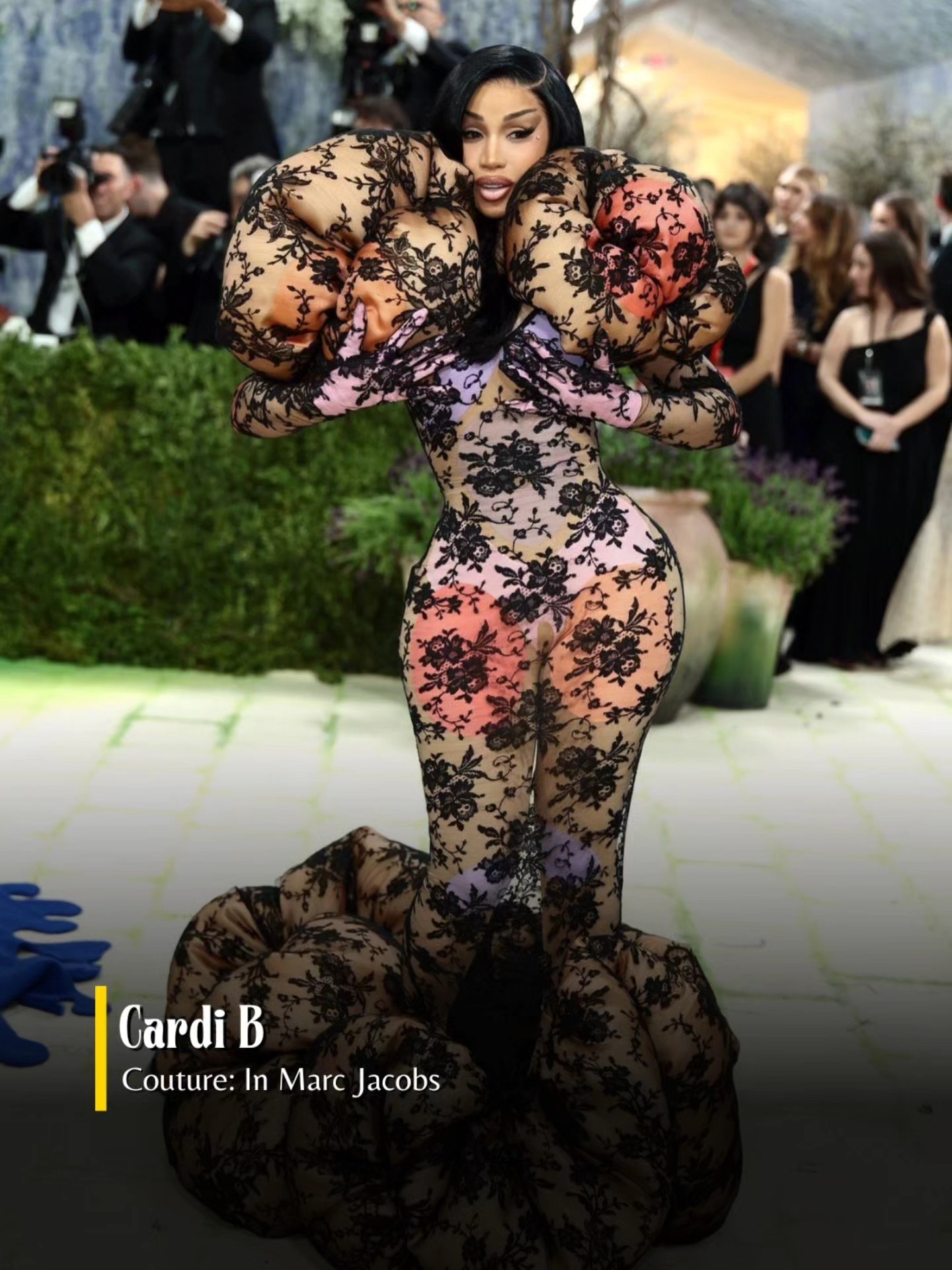 This relatable humor meme contrasts the common social expectation to dress modestly and appropriately when meeting a romantic partner's parents for the first time with the reality of someone intentionally or unintentionally wearing an extremely bold, attention-grabbing outfit. The visual used is a photo of American rapper Cardi B posing at the 2024 Met Gala red carpet, wearing an elaborate, form-fitting sheer Marc Jacobs couture gown with black lace floral detailing, large sculptural puffy elements around the shoulders and the base of the gown in matching lace, and pastel pink and orange gradient accents. Photographers and other event attendees are visible in the background of the red carpet shot, highlighting how high-profile and dramatic the outfit is, which exaggerates the joke of ignoring the dress code advice for meeting family.