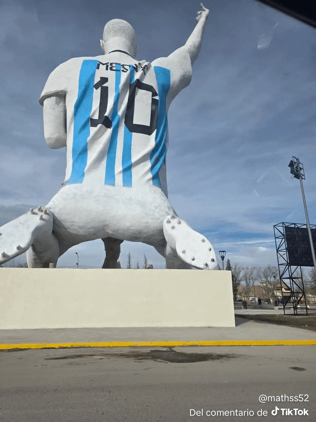 This image shows a low-angle rear view of a large, awkwardly proportioned statue created to honor famous Argentine footballer Lionel Messi, recently unveiled in Patagonia, Argentina. The statue wears a white and light blue striped Argentina national football jersey with the number 10 printed on the back, though the name above the number is visibly misspelled. The statue has very odd, disproportional dimensions, particularly its squat lower half and cleated feet that look especially absurd from this low angle, positioned on top of a plain beige concrete plinth. The background features a partly cloudy blue sky, stadium lighting poles, a digital scoreboard structure, and bare trees, confirming the statue is located in an outdoor public sports area. The humor of the meme comes from the extremely poor craftsmanship and inaccuracy of the statue meant to celebrate one of the most famous footballers in the world, making it look ridiculous and unintentionally funny.