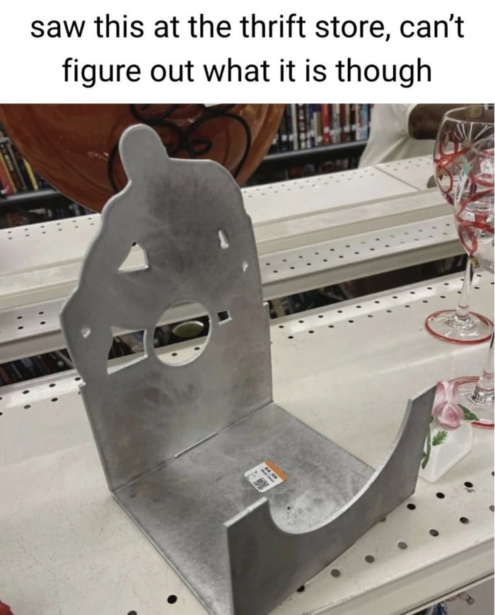 The image features a top caption stating 'saw this at the thrift store, can’t figure out what it is though' above a photograph of a mysterious silver metal object. The object, placed on a white perforated shelf in a thrift store, has an irregular, abstract shape with various cutouts, including a circular hole and triangular notches. In the background, there are shelves lined with books and decorative glassware, such as a wine glass with red patterns. A small price tag is visible on the metal object, indicating it is for sale. The scene suggests a casual thrift store environment where the poster encountered an unidentifiable item.