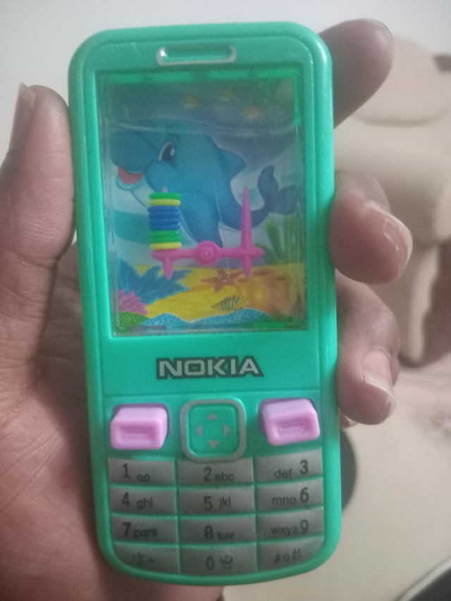 This image is a photo of a real human hand holding a cheap green plastic toy designed to look like an old Nokia feature phone. Instead of a real phone screen, the 'screen' area holds a classic children's handheld water ring toss game, filled with water, with stacked rings on a pink plastic piece. The toy has a printed Nokia logo on its front, and an underwater scene with a cartoon blue dolphin printed as the background of the game. The original poster's title indicates they have finally achieved the goal of the game: getting all the rings onto the posts.