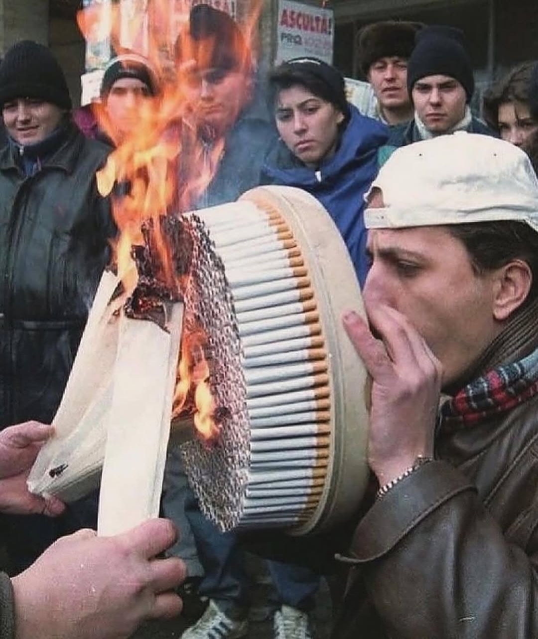 The image is a humorous digitally edited scene showing an impossibly large pack containing hundreds of standard cigarettes, held open and lit on fire at one end by a pair of hands in the foreground. A man wearing a backwards white baseball cap, leather jacket, and plaid scarf stands nearby, holding his hand near his mouth as if preparing to take an extremely large drag from the giant burning pack. A crowd of people dressed in cold weather winter clothing stand in the background watching the stunt. The comedic effect comes from the extreme, absurd exaggeration of the act of smoking, taking a common everyday activity to an unrealistic, over-the-top scale.