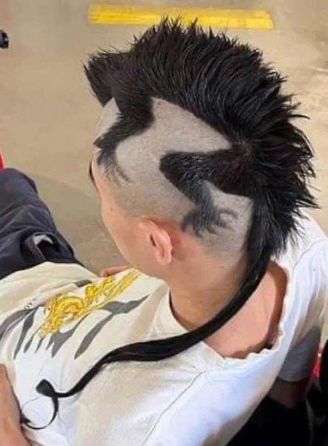 The image shows the back and side of a person's head with a novelty haircut designed to look exactly like a medical syringe. The spiky dark hair on top forms the plunger of the syringe, the shaved side of the head acts as the barrel of the syringe, and a long thin dark braid of hair trailing down the person's neck forms the syringe needle. The person is wearing a white t-shirt with a gold embroidered dragon graphic on the back, and is facing away from the camera. The humor comes from the visual pun of the syringe haircut paired with the title referencing morphine, a medication administered via syringe.
