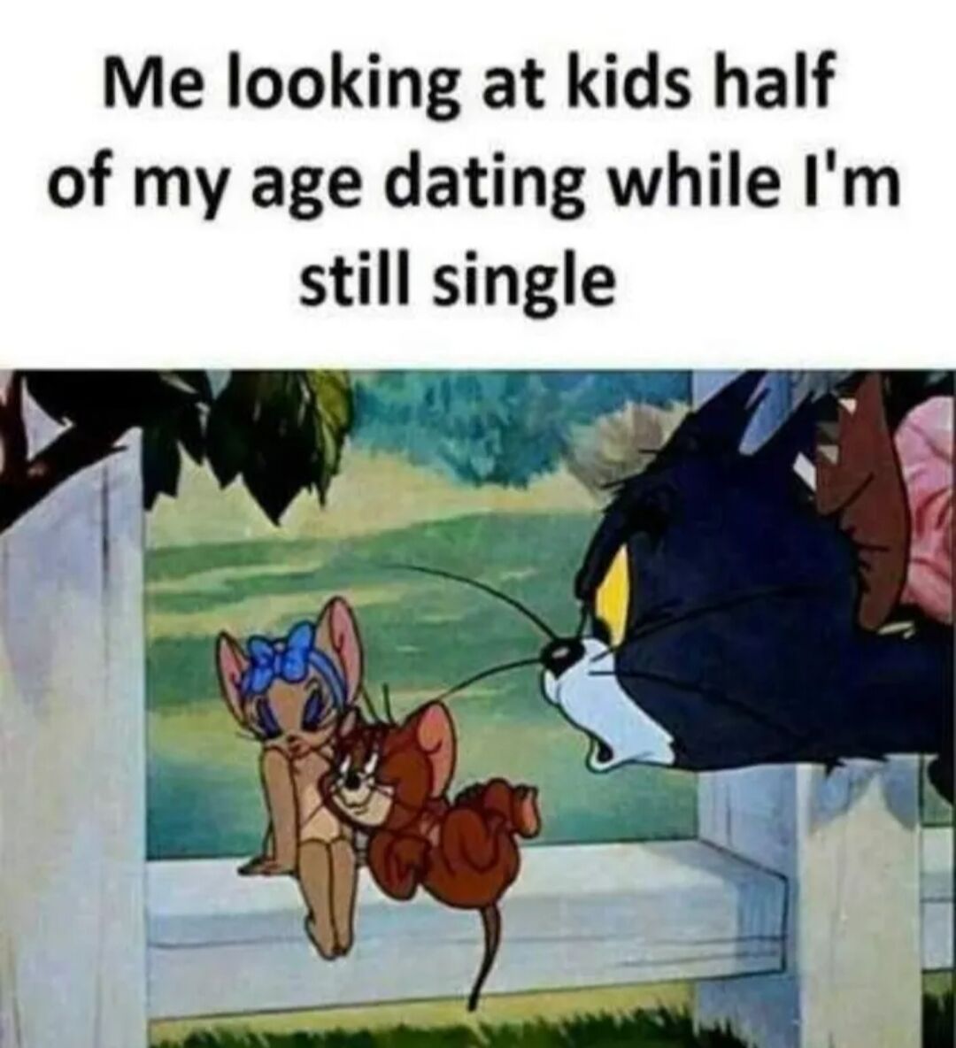 A meme featuring a top text overlay that reads 'Me looking at kids half of my age dating while I'm still single' paired with a scene from the 'Tom and Jerry' cartoon. In the scene, Tom the cat is peeking over a white fence with an envious expression, watching Jerry the mouse affectionately hugging a female mouse with a blue bow (Toots) in a grassy field. The meme humorously conveys the relatable feeling of being single while observing younger people in relationships.