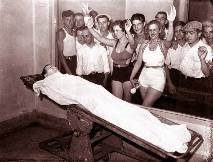A vintage black and white photograph showing a person lying on a medical examination table covered with a white sheet, surrounded by a group of approximately 10-12 men and women. The women are wearing vintage swimwear (bathing suits) while the men wear a mix of casual clothing including tank tops, button-down shirts, and one cap. Some individuals in the group have their hands raised in what appears to be an unusual gesture for a medical setting. The person on the table appears to be lying still with their head tilted back. The setting is a small room with plain walls and a mirror on one wall reflecting part of the group.