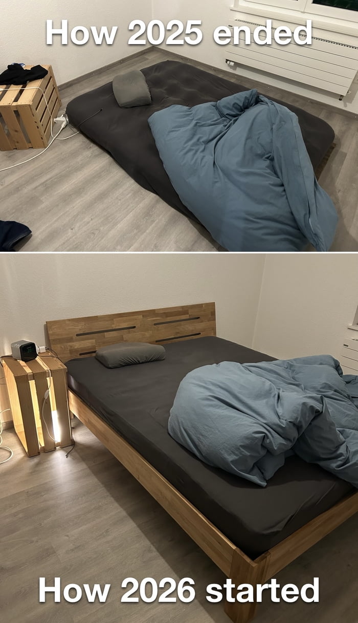 A two-panel vertical comparison meme depicting the transition from 2025 to 2026. The top panel, labeled 'How 2025 ended,' shows a basic living setup with a mattress directly on the floor, covered by a rumpled blue blanket, a gray pillow, and a wooden crate serving as a nightstand with visible cables. The bottom panel, labeled 'How 2026 started,' presents an upgraded version: the same mattress is now placed on a wooden bed frame, the crate is repurposed as a functional nightstand with a lit lamp, and the overall arrangement appears more organized and comfortable, humorously highlighting a small but positive improvement in living conditions to mark the new year.