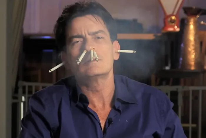 A photograph of Charlie Sheen edited to feature multiple lit cigarettes inserted in his nose and ears, with visible smoke emanating from them. He has a serious expression with furrowed brows, looking directly at the camera. The background includes indistinct household objects, notably a copper-colored kettle. The meme humorously implies that despite his controversial public image, Charlie Sheen is not associated with the Epstein files, leveraging his past reputation for excessive behavior to create a sarcastic joke.