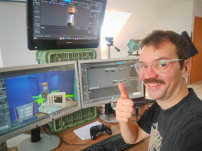 A smiling real man with glasses and a mustache gives a thumbs-up, seated at a cluttered home desk serving as a 3D artist's workspace. Three monitors display 3D creation software interfaces: the top Sony monitor shows a stylized 3D character model, while the two lower monitors display a 3D room environment and a node-based animation/editing panel (with the text 'TWEENPEAKER' visible on one). Notably, the monitors are propped up on repurposed green plastic crates instead of official stands, highlighting a budget, resourceful DIY setup. A game controller, keyboard, and desk lamp are also present on the desk, emphasizing a casual home workspace for an indie 3D creator or game developer who appears proud of their functional, low-cost setup.