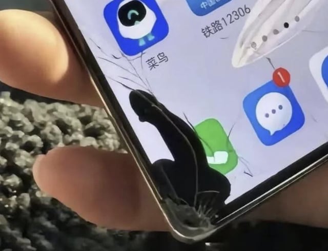 This image shows a close-up of a person holding a smartphone with a cracked screen. The pattern of the screen crack, located directly below the Cainiao logistics app icon, coincidentally forms the clear silhouette of a human figure striking a dynamic high-kick or dance pose, turning the frustrating common occurrence of a broken phone screen into a humorous visual gag. Several other common Chinese mobile apps are also visible on the home screen, including the China Railway 12306 ticketing app and WeChat, and the phone is held over a surface of loose gravel.