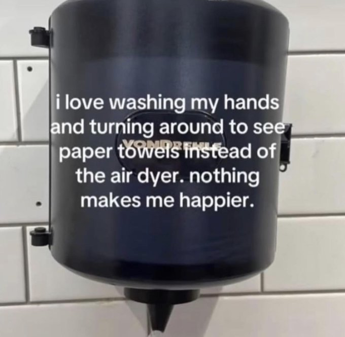 This meme features a photo of a black Vondrehle branded paper towel dispenser mounted on a white tiled public restroom wall. Overlaid on the dispenser is white text that expresses a relatable small joy: the speaker loves washing their hands in a public restroom and turning around to find paper towels available instead of an air hand dryer, stating that nothing makes them happier. The humor comes from the widely shared sentiment that public air hand dryers are often loud, slow, ineffective, and unpleasant to use, making the presence of paper towels a small but very satisfying win in daily life.