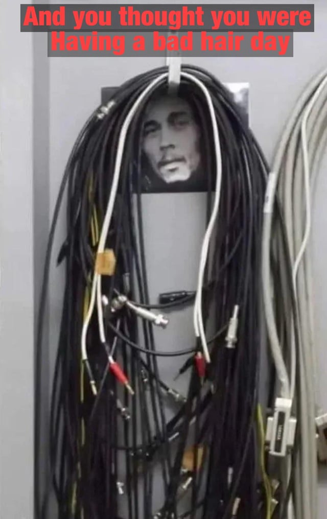 A meme featuring a tangled mess of black, white, and yellow cables hanging in front of a gray wall. Behind the cables is a black and white photograph of a man's face. The top of the image has a red caption reading 'And you thought you were having a bad hair day,' using the messy, unkempt cables as a humorous visual comparison to unruly hair on a bad hair day.