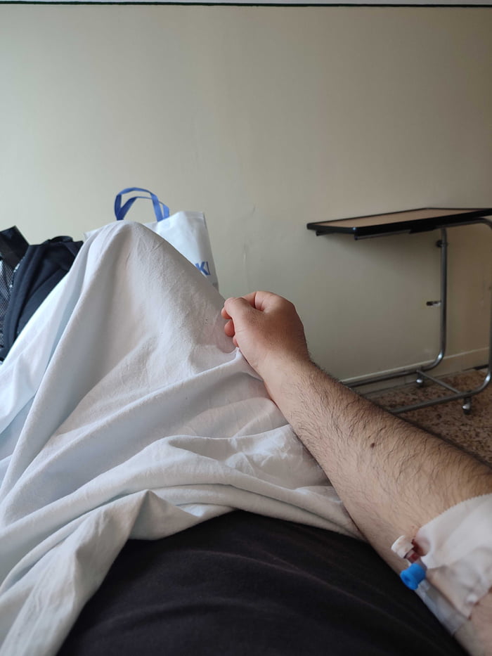 This is a first-person personal photo taken from a hospital bed, showing the poster's forearm with an intravenous (IV) cannula inserted for medical treatment, resting over a wrinkled white hospital sheet and dark clothing. In the background, there is a plain beige wall, a rolling over-bed hospital table, and personal bags behind the sheet. The accompanying post explains that the poster was admitted to hospital for a sinus infection, and unexpectedly received a brain tumor diagnosis, saying goodbye to his community.