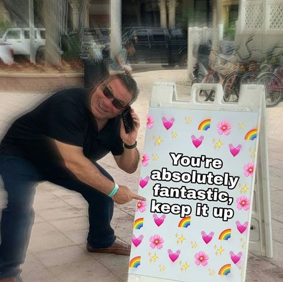 This is a remixed meme featuring actor James Gandolfini, famous for portraying the tough mob boss Tony Soprano in *The Sopranos*, in an outdoor urban setting. Gandolfini is crouched down, holding a flip phone in one hand and pointing enthusiastically at an edited A-frame sign with the other. The sign is adorned with playful, cute graphics including pink hearts, rainbows, flowers, and sparkles, and displays the warm, encouraging message: **You're absolutely fantastic, keep it up**. The core humor comes from the stark contrast between Gandolfini's iconic, intimidating on-screen persona and the gentle, uplifting message on the sign, creating a funny yet heartwarming effect. This meme is designed to be a supportive, positive reaction to someone's efforts or achievements.