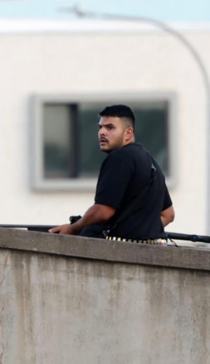 A photograph of a man with short black hair wearing a black short-sleeved shirt, leaning on a weathered concrete ledge. He is positioned with his left arm resting on the ledge, looking to his right with a serious and alert expression. Behind him, a white building with a rectangular window is visible, slightly out of focus, suggesting an urban outdoor setting. The man has a utility belt with various items around his waist, potentially indicating a security, maintenance, or similar role. The concrete ledge appears weathered, with visible texture and slight discoloration, and the sky above is a light blue, indicating daytime.