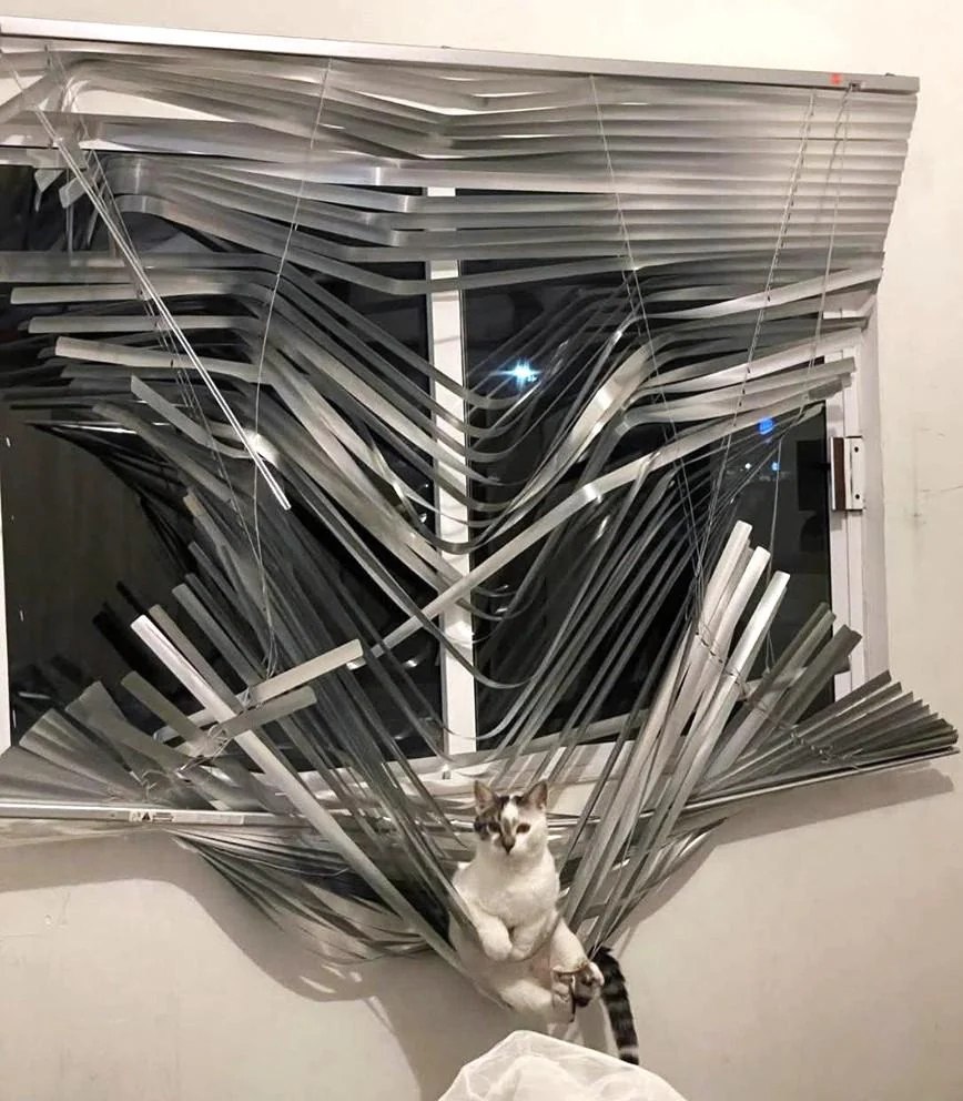 The image depicts a window with severely bent, twisted, and disarrayed aluminum blinds, appearing as if they were forcefully manipulated or damaged. In front of the chaotic blinds sits a white cat with a tabby-colored face, which looks calm and unbothered. The stark contrast between the destroyed blinds and the cat's nonchalant demeanor humorously suggests the cat may be responsible for the mischief, playing on the common trope of pets causing household destruction.