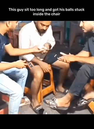 This image is a still frame from a real-life viral video. It shows three men sitting outdoors. The man in the center, wearing a white shirt, is positioned on a small low wooden stool, gripping its edges and looking down with an uncomfortable, pained expression. The two men sitting on either side of him are holding mobile phones, seemingly recording the central man's predicament. White text at the top of the image reads "This guy sit too long and got his balls stuck inside the chair", explaining the awkward, embarrassing and painful situation the central man is in. The humor of the meme comes from the absurd, relatable awkwardness of the embarrassing predicament and the casual, amused reaction of the bystanders recording the incident.