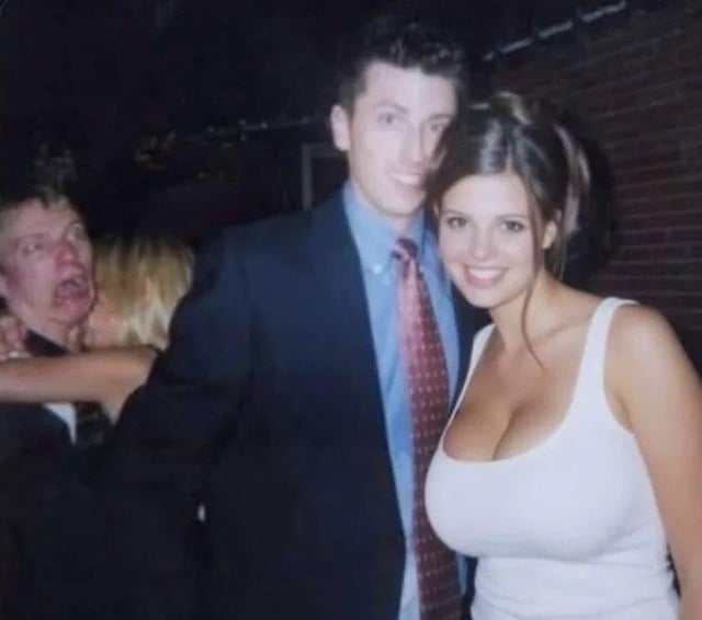 This is a low-light party photograph meme. The foreground features a smiling woman in a form-fitting white tank top standing next to a smiling man in a dark suit, light blue shirt, and patterned red tie, both posing directly for the camera to draw the viewer's immediate attention. In the far left background, a pale man with an exaggerated wide open mouth and wide eyes (jokingly referred to as a vampire) stands while a blonde woman appears to be kissing or biting his neck. The core joke of the meme is that the attractive woman in the foreground acts as a decoy to distract viewers from noticing the bizarre, humorous vampire-like figure and scene in the background right away.
